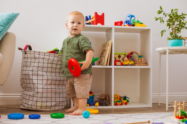 Der kleine blonde Junge holt Spielzeug aus der Tasche Er steht mit einem Spielzeug im Spielzimmer und sieht sich um