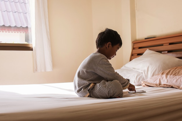 Der kleine Junge, der auf Bett sitzt, spielt intelligentes Telefon im Schlafzimmer