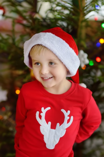 Der kleine Junge, der den Sankt-Hut betriebsbereit ist, feiern Weihnachten. Nettes Kind, das den Weihnachtsbaum mit Glasspielzeug verziert