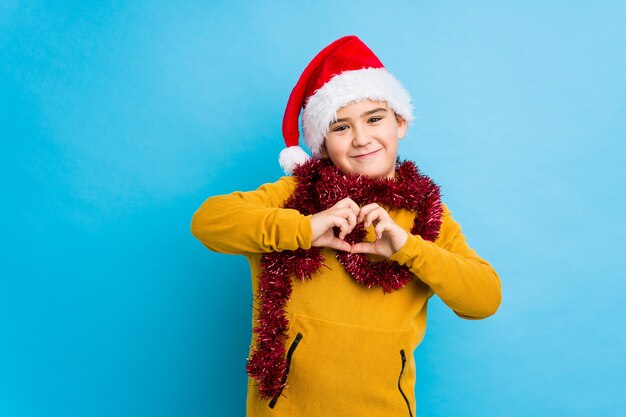 Der kleine Junge, der den Weihnachtstag trägt einen Sankt-Hut feiert, lokalisierte das Lächeln und das Zeigen einer Herzform mit den Händen.