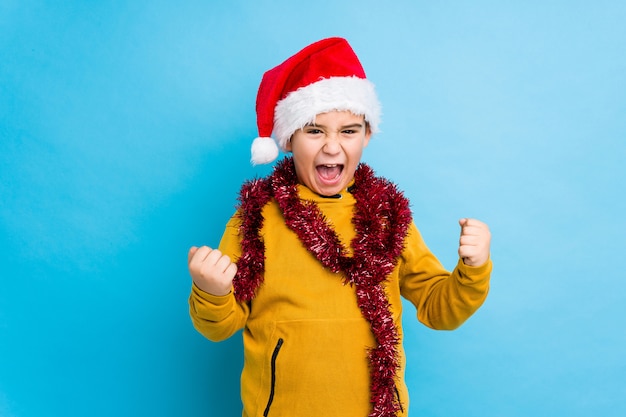 Der kleine Junge, der den Weihnachtstag trägt einen Sankt-Hut feiert, lokalisierte das Zujubeln sorglos und aufgeregt. Sieg-Konzept.