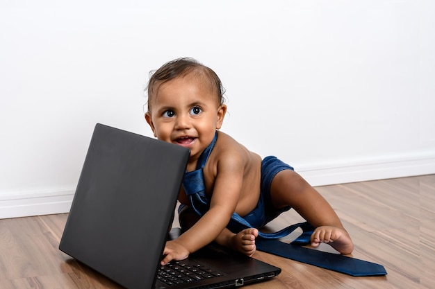 Der kleine Junge, der mit dem Laptop spielt, schaut in die Kamera, trägt eine Krawatte und sitzt ohne Hemd auf dem Holzboden