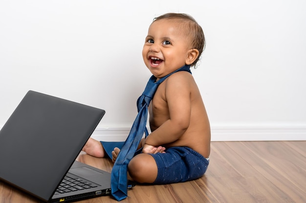 Der kleine Junge, der mit dem Laptop spielt, schaut in die Kamera, trägt eine Krawatte und sitzt ohne Hemd auf dem Holzboden