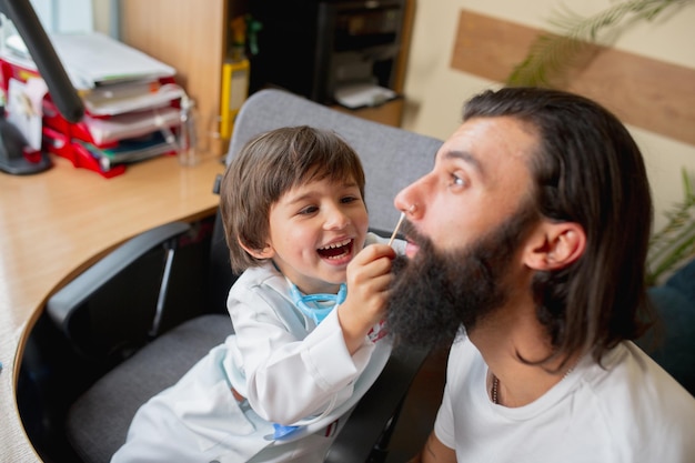 Der kleine Junge, der spielt, tut so, als ob ein Arzt einen Mann in einer komfortablen Arztpraxis untersucht