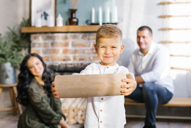 Der kleine Junge hält einen Baumstamm in den Händen, um vor dem Hintergrund glücklicher Eltern einen Kamin anzuzünden