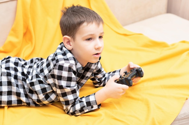 Der kleine Junge im karierten Hemd liegt auf der Couch mit schwarzem Joystick in den Händen und spielt das Videospiel Videospiele zu Hause spielen Konzept