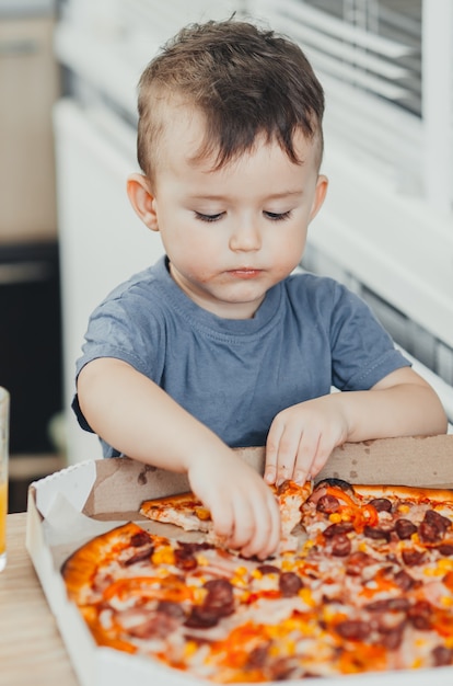 Der kleine Junge isst selbst in der Küche eine riesige schädliche Pizza und trinkt Saft, sehr fett und schädlich