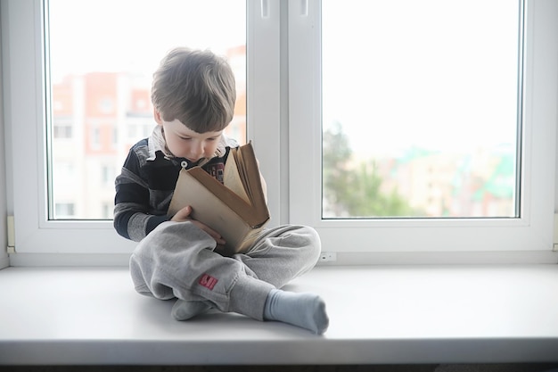 Der kleine Junge liest ein Buch Das Kind sitzt am Fenster und bereitet sich auf den Unterricht vor Auf der Fensterbank sitzt ein Junge mit einem Buch in der Hand