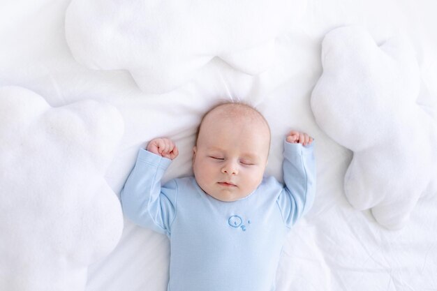 Der kleine Junge schläft auf dem Bett, das auf dem Rücken liegt, in einem blauen Pyjama, mit den Händen zwischen den Kissen der Wolken, gesunder Neugeborenenschlaf