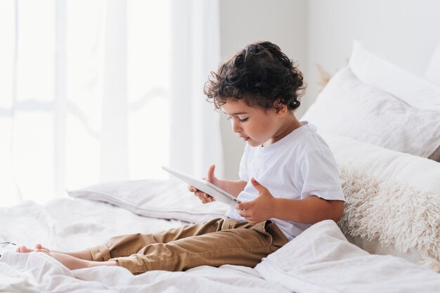 Der kleine lockige kaukasische Junge sitzt morgens mit einem Tablet auf dem Bett zu Hause mit leerem Platz im Hintergrund. Der hübsche brasilianische Kindervorschüler lernt Buchstaben über das Internet und bereitet sich auf die Schule vor