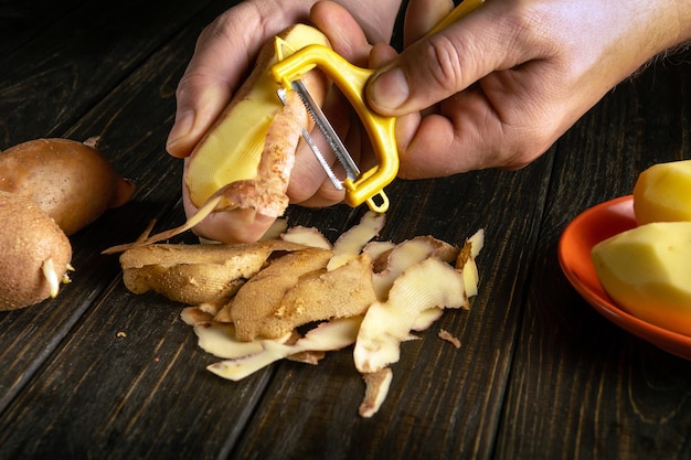 Der Koch schält Kartoffeln zum Grillen oder zur Zubereitung von Kartoffelgerichten Auf einem schwarzen Tisch Nahaufnahme von einem Koch Hände mit einem Kartoffel-Schäler