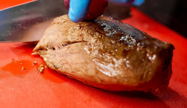 Der Koch schneidet ein saftiges gebratenes Stück Rindfleisch mit einem scharfen Messer, das seine Hand in einem Handschuh hält. Foto in hoher Qualität