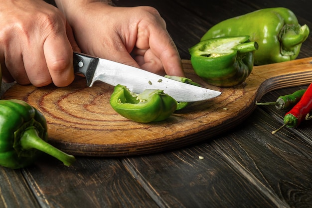 Der Koch schneidet frische grüne Paprika auf einem Holzschneidebrett Nahaufnahme der Hände des Küchenchefs bei der Zubereitung vegetarischer Speisen