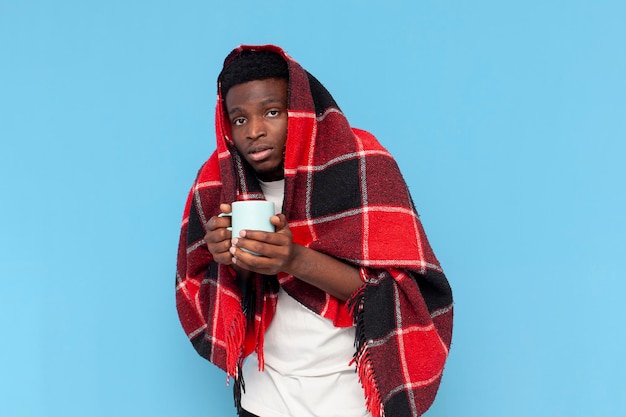 Der kranke junge Afroamerikaner, der etwas trinkt, leidet unter Fieber und ist mit warmem Plaid bedeckt