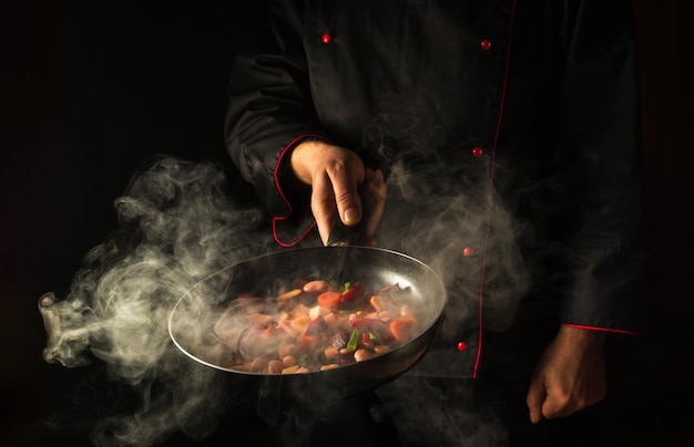 Der Küchenchef bereitet Speisen in einer heißen Pfanne mit Dampf auf schwarzem Hintergrund zu
