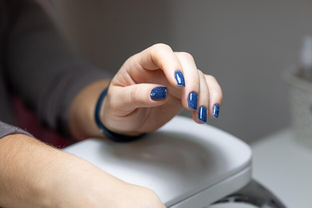 Der Kunde hält seine Hand auf einem Ständer mit einem blauen Gellack, der auf die Nägel aufgetragen wird