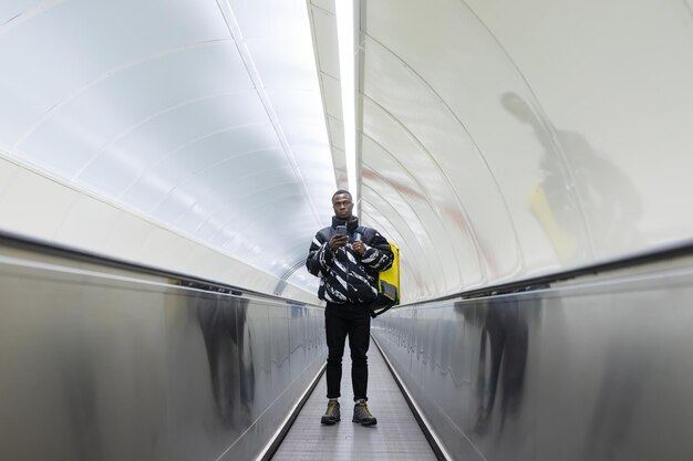 Der Kurier fährt mit einem Handy und einem gelben Rucksack auf der Rolltreppe Lebensmittellieferung aus dem Laden