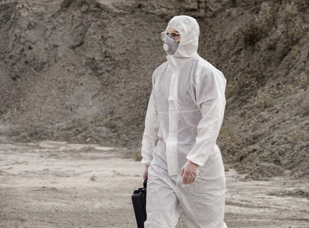 Der Labortechniker in Maske und Chemikalienschutzanzug geht mit einem Werkzeugkasten auf trockenem Boden durch giftigen Rauch.