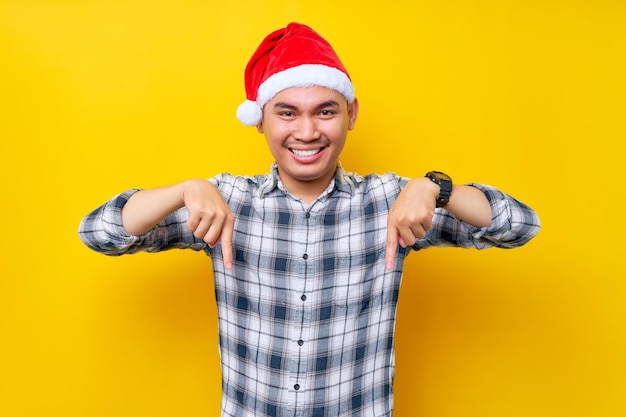 Der lächelnde junge asiatische Mann, der ein kariertes Hemd in einer Weihnachtsmütze trägt und mit dem Finger nach unten zeigt, zeigt den Arbeitsbereich auf gelbem Hintergrund an, um die Weihnachtsfeiertage und das Neujahrskonzept zu feiern