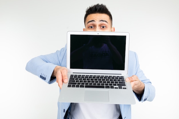Der lächelnde junge brünette Mann hält freudig einen Laptop mit Leerzeichen zum Einfügen einer Webseite auf einem weißen Hintergrund.