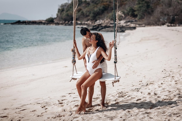 Der liebevolle Ehemann küsst sanft seine schwangere Frau und umarmt ihren Bauch, während sie auf den Schaukeln am Sandstrand am Wasser sitzt; Harmonie-Konzept.