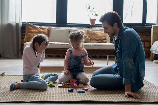 Der liebevolle junge Vater, der mit kleinen Kindern auf dem Boden liegt, hat Spaß an lustigen Aktivitäten mit Bauziegeln, fürsorglicher Vater spielt mit kleinen Kindern, die mit Blöcken bauen, genießen die Zeit zu Hause zusammen