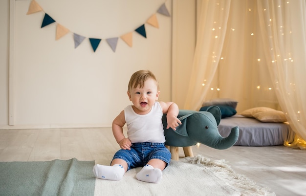 Der lustige kleine Junge in einem weißen Body und Jeans sitzt mit Spielzeug auf dem Boden im Kinderzimmer und schaut in die Kamera