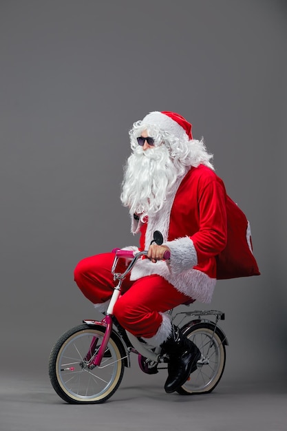 Der lustige Weihnachtsmann mit Sonnenbrille und Weihnachtsgeschenken auf dem Rücken fährt Fahrrad