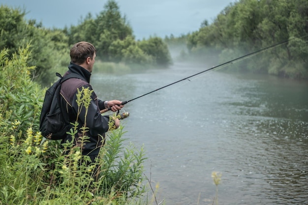 Der männliche Fischer steht mit einer Angelrute in den Händen am Flussufer und fängt Fische Ruhige Erholung im Freien