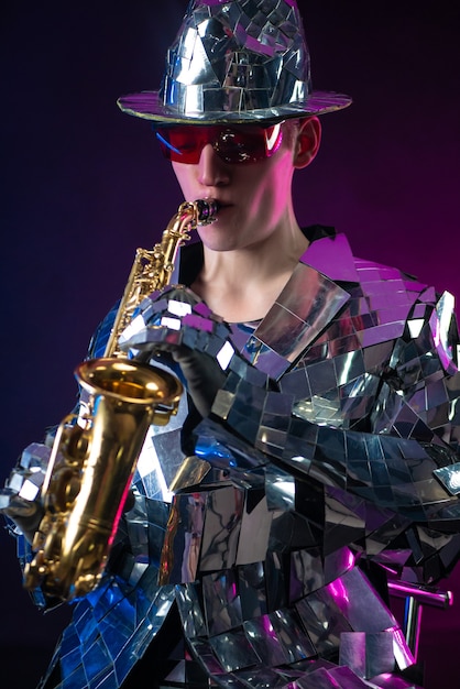 Der männliche Künstler im verspiegelten Anzug und Hut mit Saxophon im Neonlicht