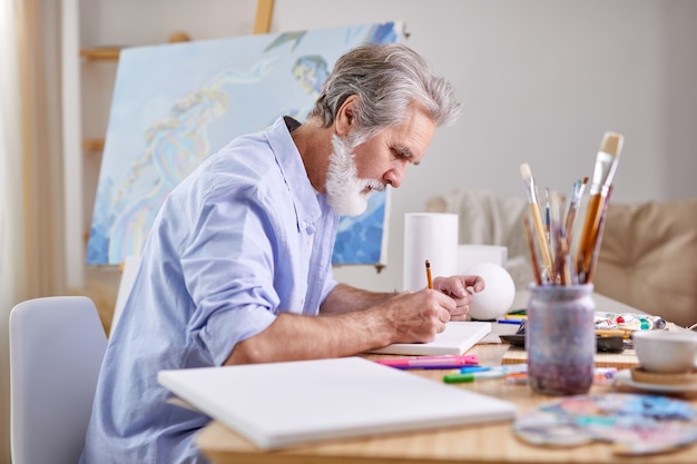 Der Maler zeichnet zu Hause mit Bleistift, konzentriert sich auf die Arbeit und trägt ein blaues Hemd.