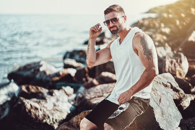 Der Mann am Erholungsort in einem weißen Hemd und in einer Sonnenbrille, die auf einem Felsen auf dem Meer sitzen