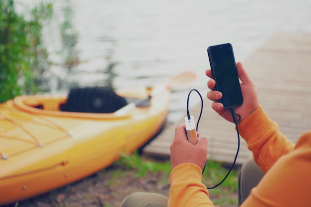 Der Mann hält ein tragbares Ladegerät mit einem Smartphone in der Hand. Ein Mann vor dem Hintergrund von Wasser und Kajak lädt das Telefon mit der Power Bank auf.