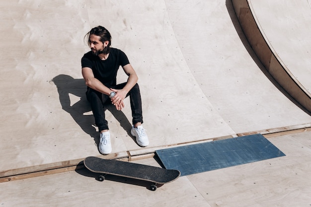 Der Mann in der stylischen Freizeitkleidung sitzt am sonnigen Tag auf der Rutsche neben dem Skateboard in einem Skatepark.