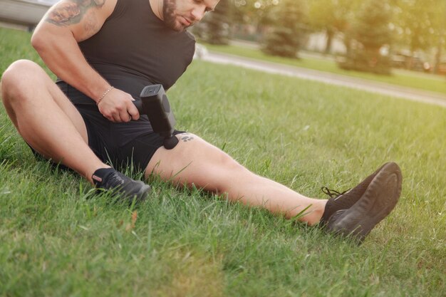 Der Mann massiert den Körper mit dem Massage-Percussion-Gun-Gerät während des aktiven Outdoor-Sports. Massagepistole zur Selbstwiederherstellung der Muskelleistung während des Trainings. Physiotherapie von Sportlern. Outdoor-Aktivitäten