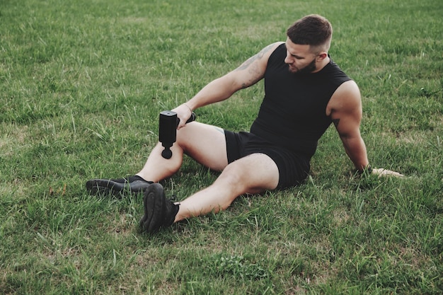 Foto der mann massiert den körper mit dem massage-percussion-gun-gerät während des aktiven outdoor-sports. massagepistole zur selbstwiederherstellung der muskelleistung während des trainings. physiotherapie von sportlern. outdoor-aktivitäten