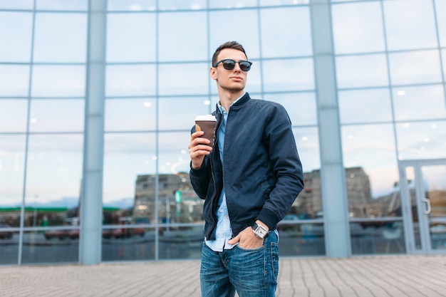 Der Mann mit der stilvollen Sonnenbrille, ein Mann geht durch die Stadt und trinkt Kaffee aus einer Pappbecher, ein gutaussehender Mann geht herum und ruht sich aus, der Mann auf dem Hintergrund eines modernen Gebäudes, Büro