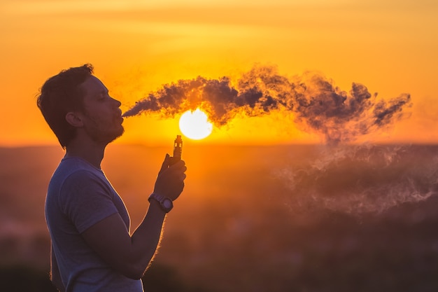 Der Mann raucht eine elektrische Zigarette auf dem Hintergrund des Sonnenuntergangs