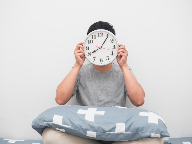 Der Mann sitzt auf dem Bett und hält die analoge Uhr in der Nähe seines Gesichts