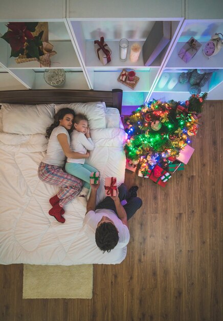 Der Mann sitzt mit Geschenken neben seiner Familie auf dem Bett. Nacht, Ansicht von oben