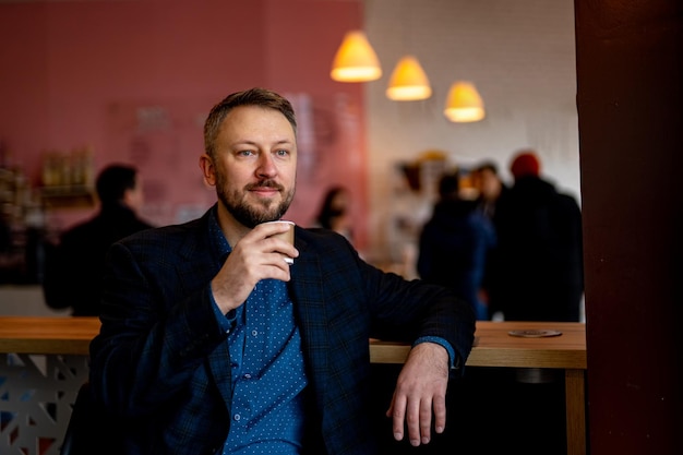 Der mann trägt einen dunklen anzug und trinkt kaffee. vorderansicht eines erfolgreichen, gutaussehenden bärtigen mannes. modernes café unscharfer hintergrund.