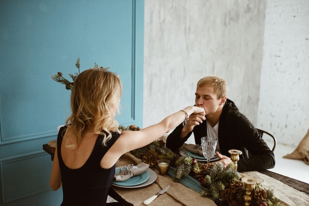 Der Mann und das Mädchen essen am festlichen Tisch zu Abend. Blond im schwarzen Kleid, mit rotem Lippenstift auf den Lippen. Besteck. Blaue Wand. Verzierter Weihnachtsbaum.