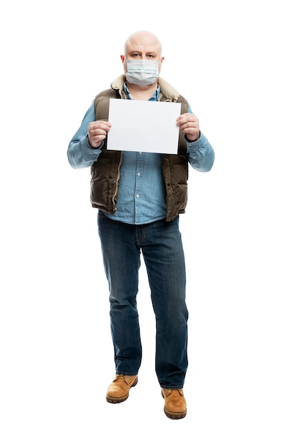 Der maskierte Mann hält ein leeres weißes Blatt Papier in der Hand. Vorsichtsmaßnahmen während der Coronavirus-Pandemie. Isoliert auf einer weißen Wand. Vertikale. Vollständige Höhe.