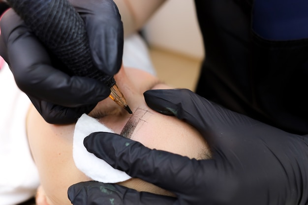 Der Meister des Permanent Make-ups führt ein Tattoo der Augenbrauen mit einer Tätowiermaschine durch, die die Augenpartie mit einem Baumwollschwamm hält