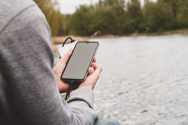 Der Mensch nutzt das Smartphone beim Laden von der Power Bank am Flussufer. Modernes Technologiekonzept Selektiver Fokus