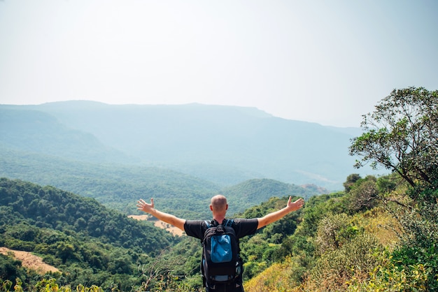 Der Mensch steht auf einem Hügel und schaut auf die Berge, die am Sanny-Tag mit Wald bedeckt sind. Brutaler Glatzkopf mit Rucksack breitete die Arme zur Seite aus, Blick von hinten. Konzept von Freiheit und Reisen