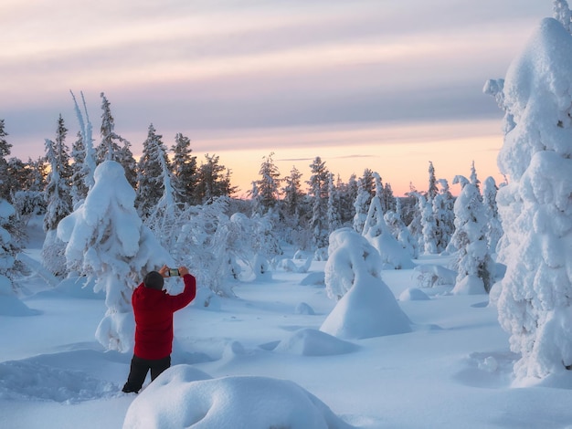 Der Mensch steht mit dem Rücken im Tiefschnee und fotografiert mit einem Smartphone die atemberaubende Aussicht auf schneebedeckte Bäume während einer kalten Polardämmerung Winterurlaub Konzeptreise in den arktischen Feenwald