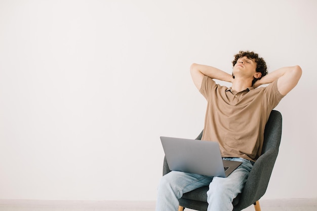 Der müde junge Mann, der einen Laptop benutzt, während er in einem Sessel vor einer weißen Wand sitzt, Kopierraum Tausendjähriger Mann, der online kommuniziert, arbeitet oder aus der Ferne auf einem tragbaren PC lernt