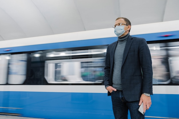 Der nachdenkliche Geschäftsmann trägt eine chirurgische sterile Maske, um die Ausbreitung von Atemwegserkrankungen zu verhindern, posiert gegen die U-Bahn, fährt mit öffentlichen Verkehrsmitteln und hält das Smartphone in der Hand. Coronavirus-Ausbruch.