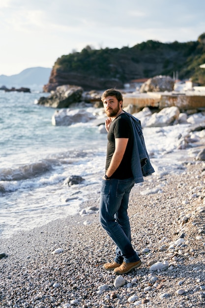 Der nachdenkliche Mann steht mit einer Jeansjacke über der Schulter an einem Kieselstrand